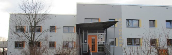 Zu sehen ist ein renovierter Plattenbau. Die Farben sind Grau und orange. Das Gebäude besitzt drei Geschosse und einen großen Eingangsbereich.